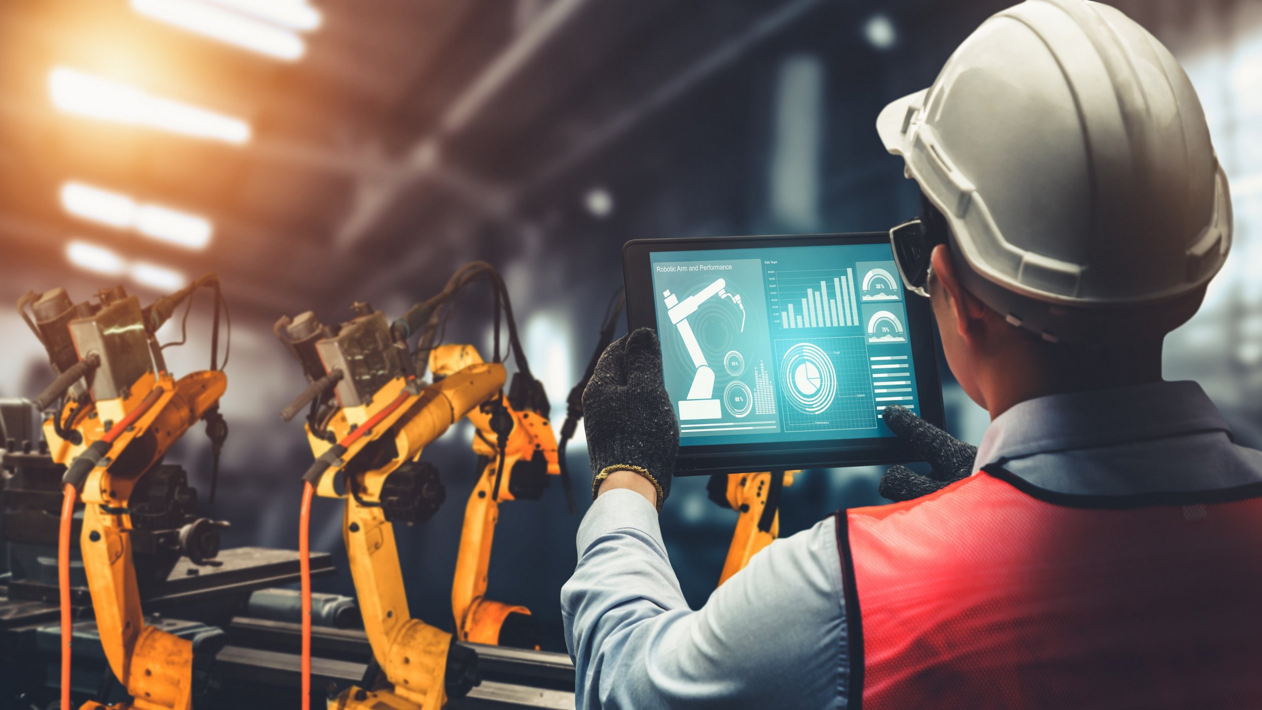 Employee wearing PPE looking at charts on a tablet in a factory with robotic equipment