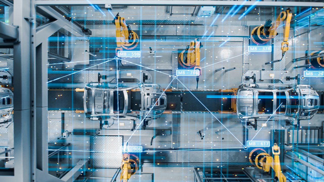 An overhead view of a car frame moving down an assembly line with industrial robot arms along the side of it and surrounded by graphics of blue data passing through the factory. 