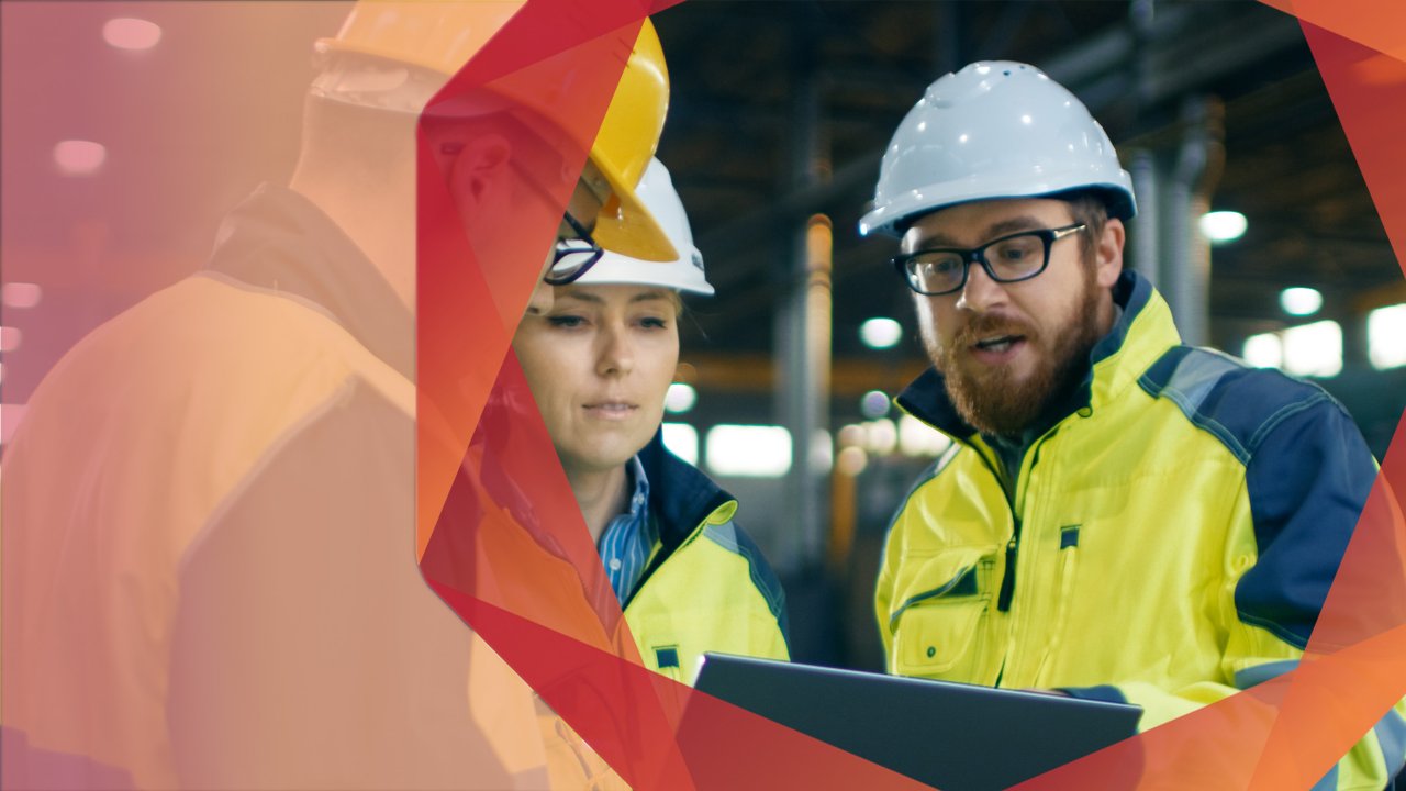 Man and woman wearing white hard hats, yellow safety jackets and protective eye wear looking at a tablet in an industrial setting.