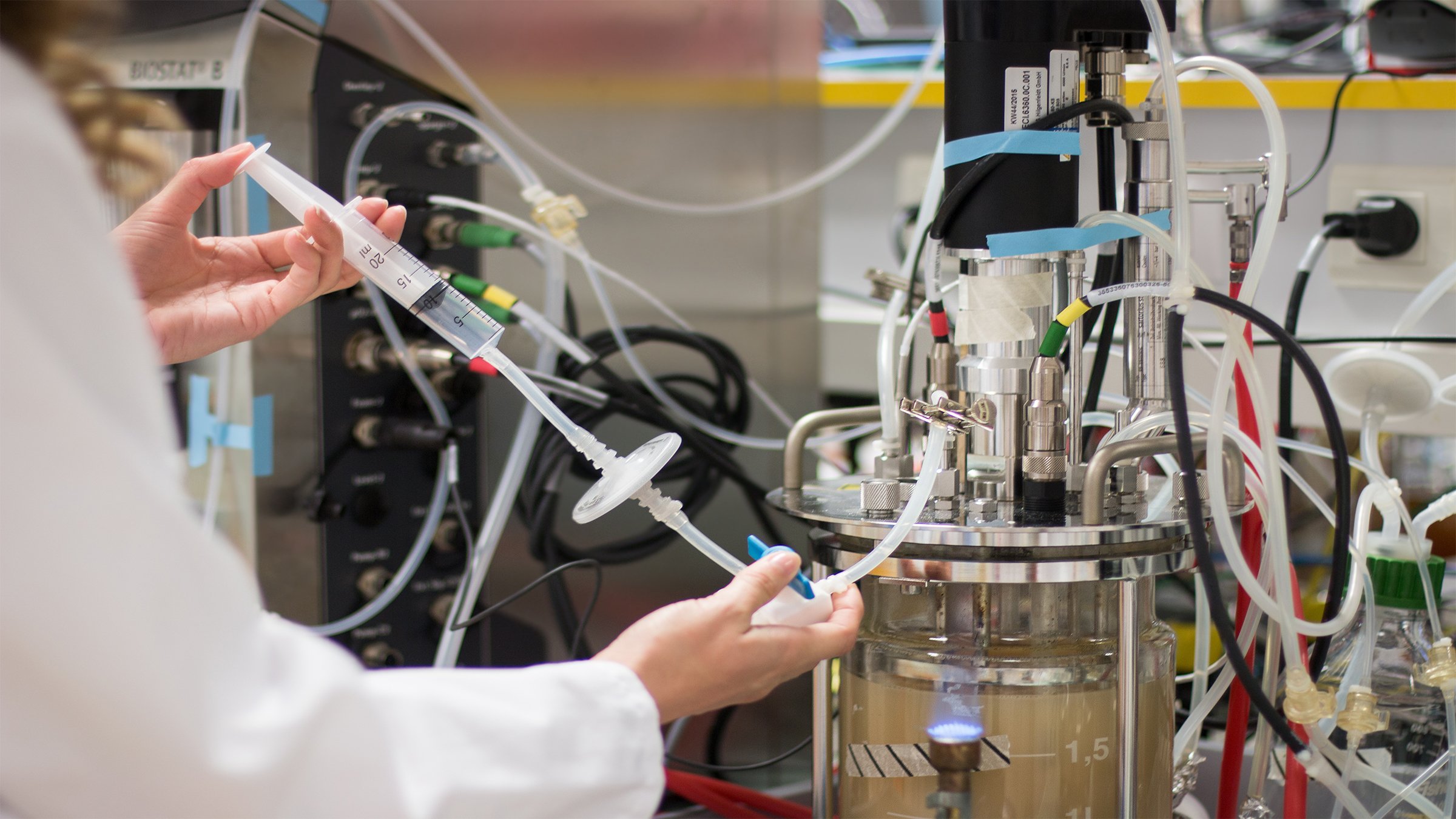 一位生命科学专业人员身穿个人防护装备，使用一次性设备将溶液注入生物反应器里。