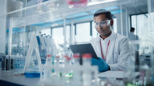 Medical Research Laboratory: Portrait of a Handsome Male Scientist Using Digital Tablet Computer to Analyse Data. Advanced Scientific Lab for Medicine, Biotechnology, Microbiology Development. Test tubes and equipment are on the table around him.