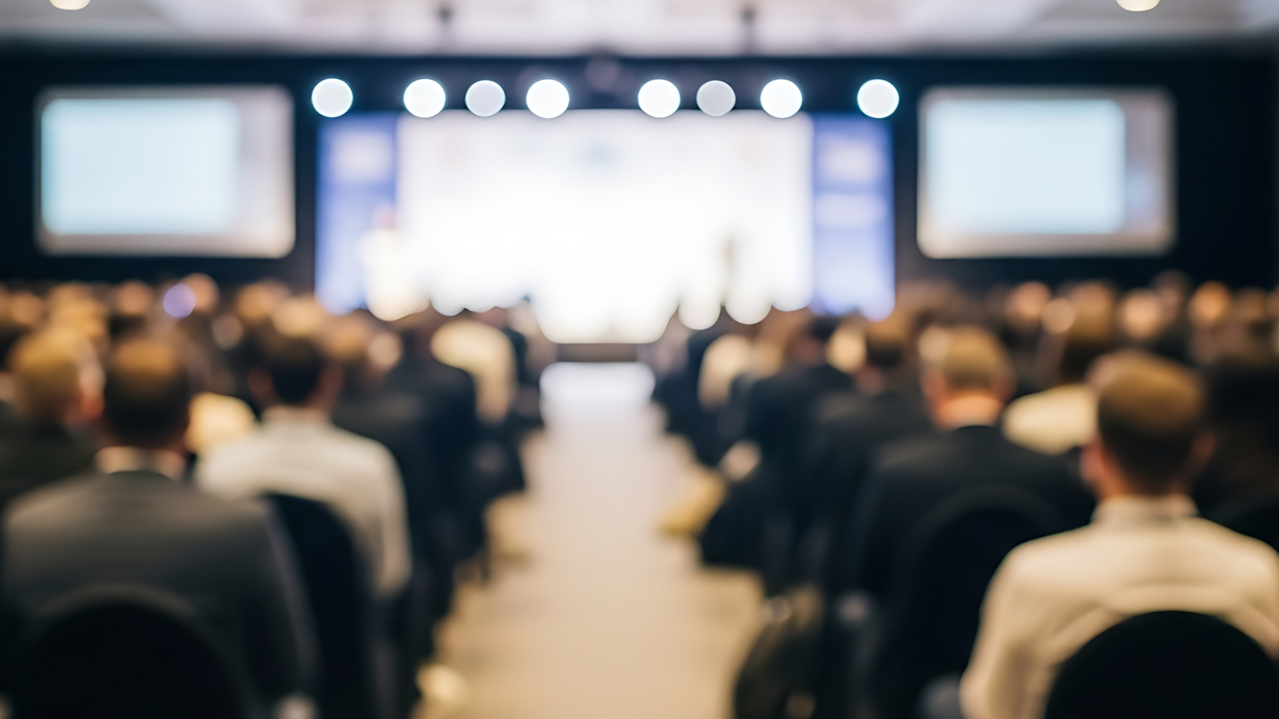 A wide-angle, out-of-focus shot taken from the back of a large conference hall, showing an audience seated in rows facing a brightly lit stage. The lighting is soft and ambient, creating a bokeh effect throughout the room, with two large projection screens visible on either side of the stage area. The atmosphere suggests a formal business seminar, corporate presentation, or an industry summit where attendees are listening to a speaker.