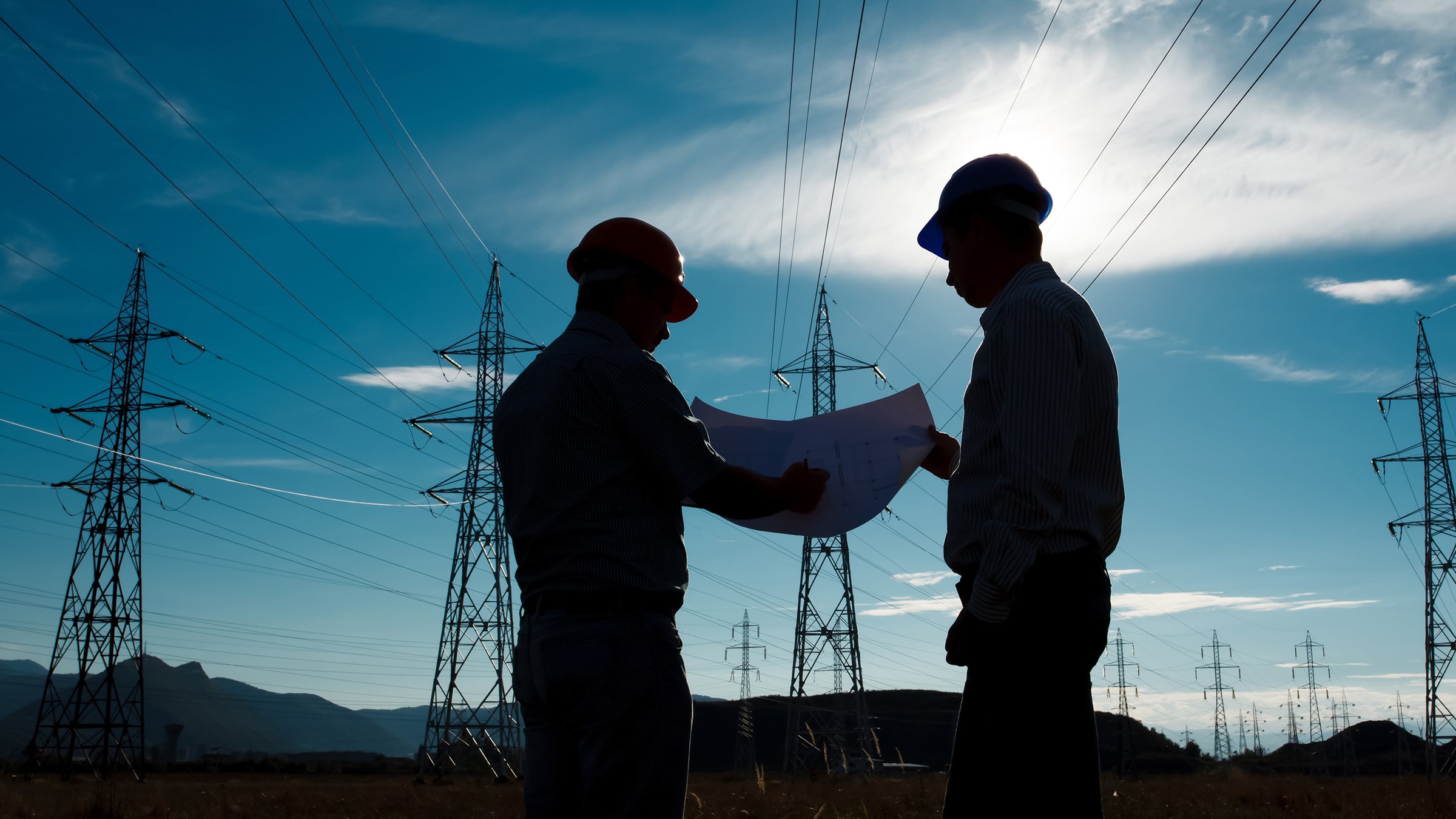 Silhouette of two male engineers in hard hats standing at electricity station, discussing critical infrastructure and how to protect it from hackers.