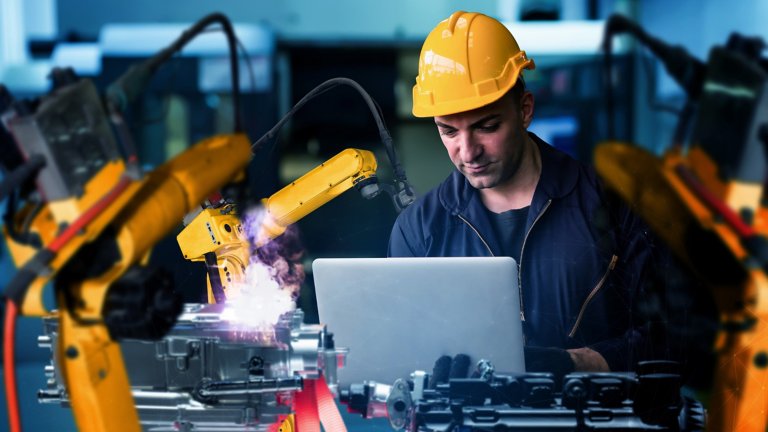 Rockwell-Techniker mit Schutzhelm und Laptop, der in einem modernen Fertigungsbetrieb mit Industrierobotern arbeitet – symbolische Darstellung des IIoT-Konzepts