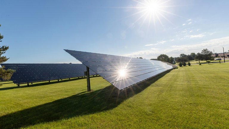 Solar panels in a field of green grass with the sun shining brightly and its reflection bouncing off one of the panels.