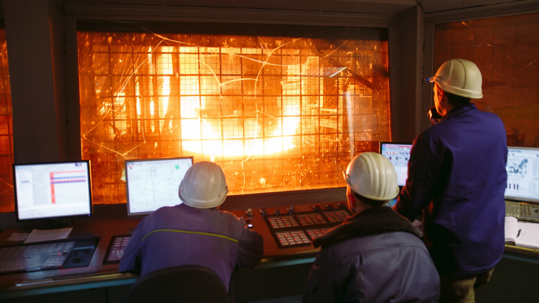 Multiple steel workers in plant control room looking through window to blast furnace 