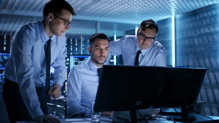 Three male engineers looking at desktop computer with two screens in industrial cybersecurity data center