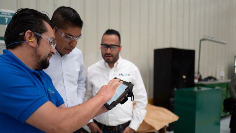 Três profissionais de operações em pé no chão de fábrica, usando óculos de segurança e visualizando os painéis do Plex Smart Manufacturing em um tablet.