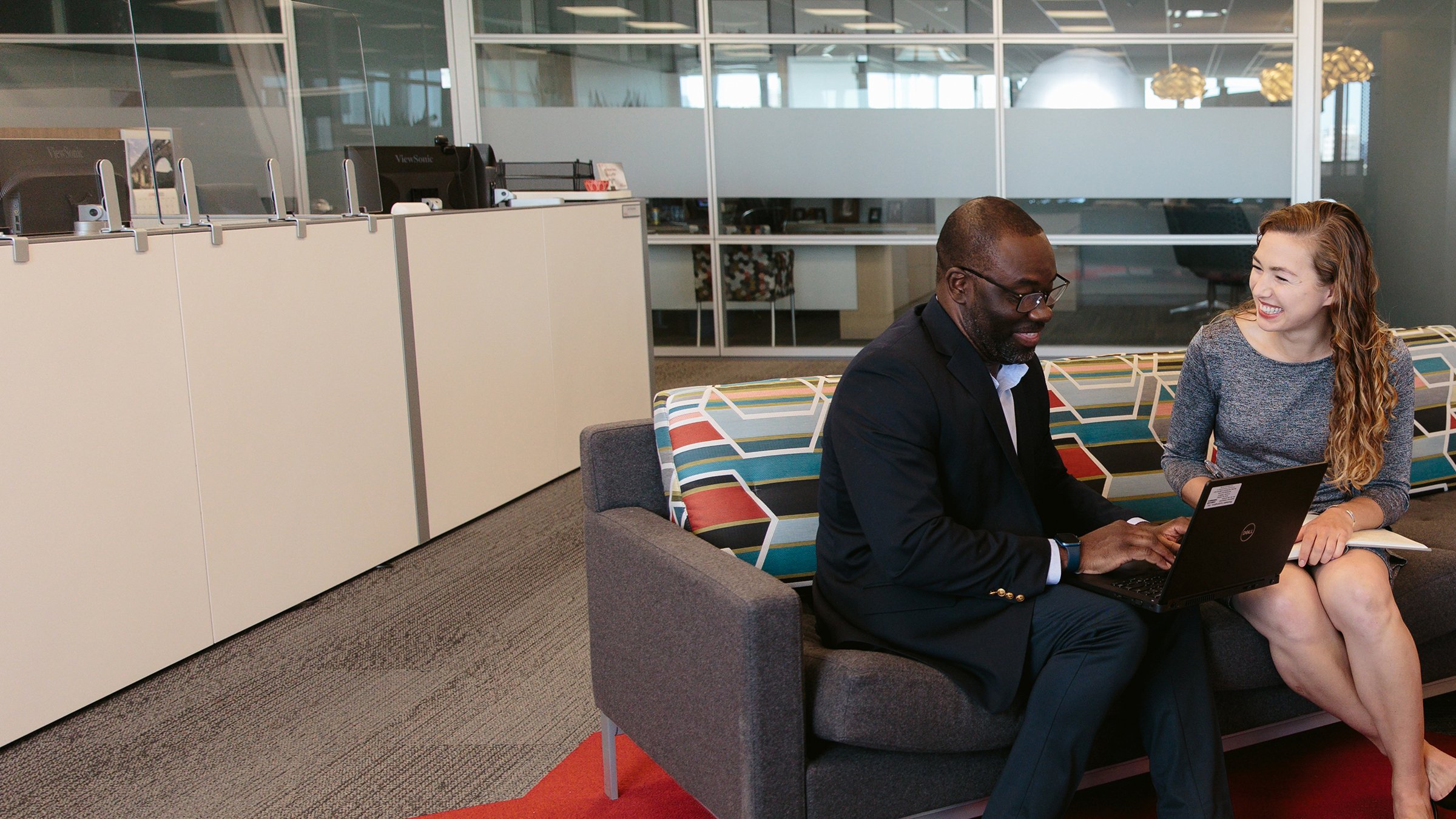 Two diverse employees meet at Rockwell Automation Headquarters. They are sitting on a couch and smiling and looking at a laptop. 