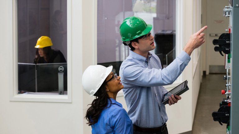 一男一女两位工程师戴着安全帽，正在检查高大的灰色工业橱柜，该橱柜具有多个门、按钮、触摸屏界面和闩锁。CENTERLINE 2100 电机控制中心。