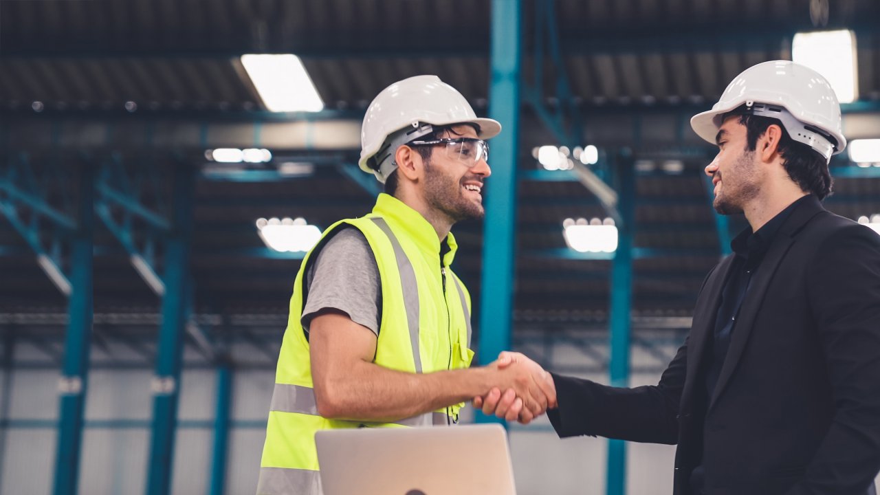  Two factory workers in hardhats standing and shaking hands in front of laptop in plant nd discussing manufacturing plan in the factory . Industry and engineering concept