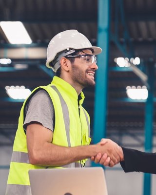  Two factory workers in hardhats standing and shaking hands in front of laptop in plant nd discussing manufacturing plan in the factory . Industry and engineering concept