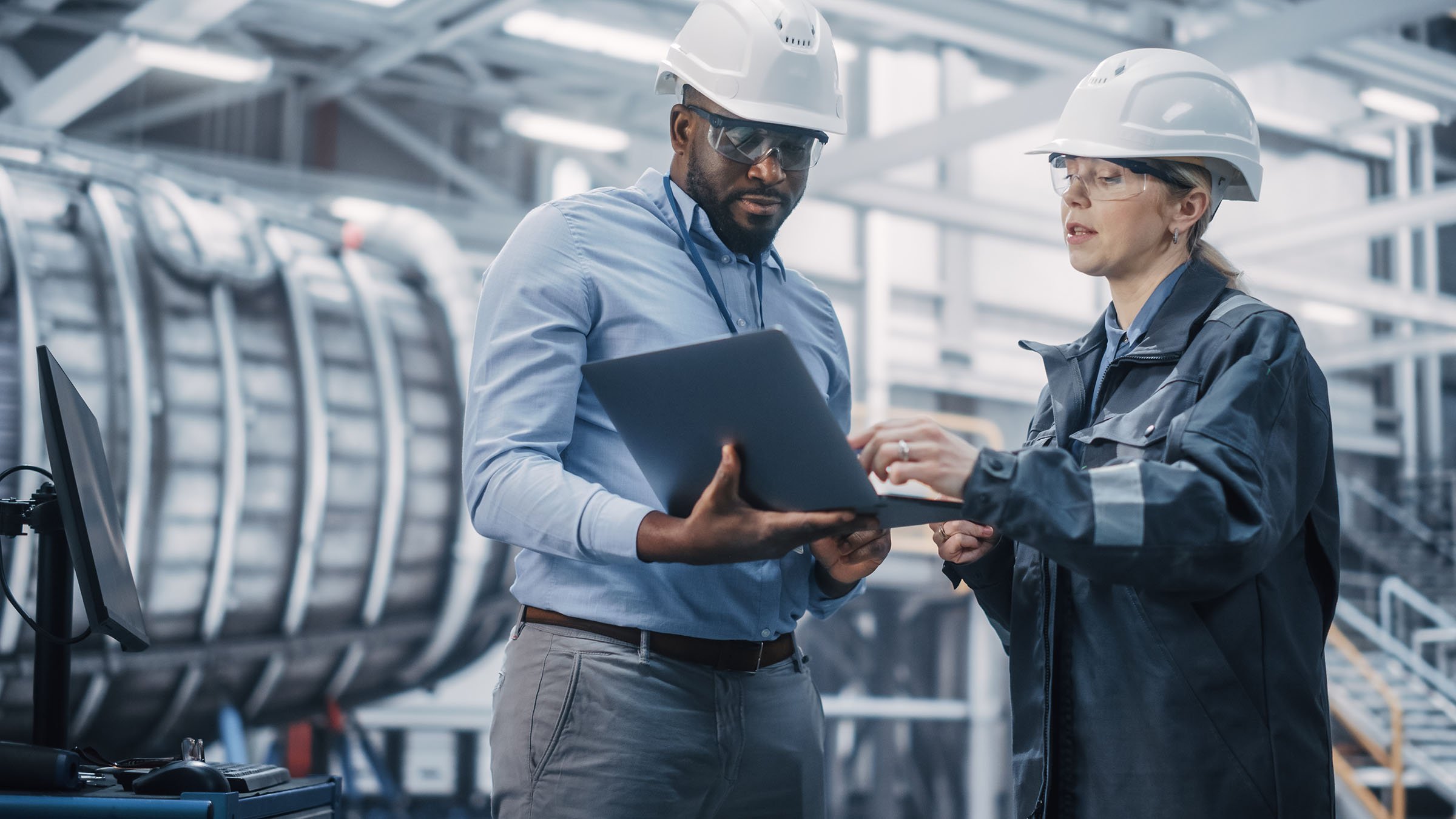 Two heavy-industry engineers in a factory wearing safety apparel working on a laptop.