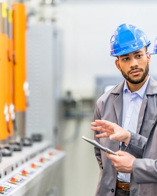 Two men in blue hard hats holding a tablet have a discussion by a machine on the plant floor