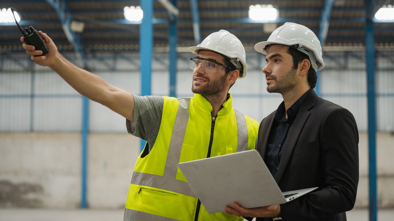 Two male factory workers in safety gear. One is holding a laptop the other has a communication device. They are looking up at something. They are in a large open building area.