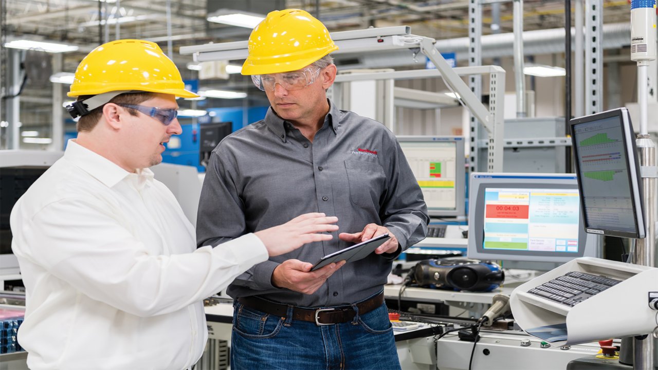 Dois homens com capacetes amarelos e óculos de segurança conversando em um ambiente industrial com um tablet na mão