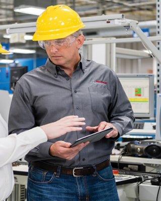 Two men in yellow hard hats and safety glasses in discussion holding tablet in an industrial environment