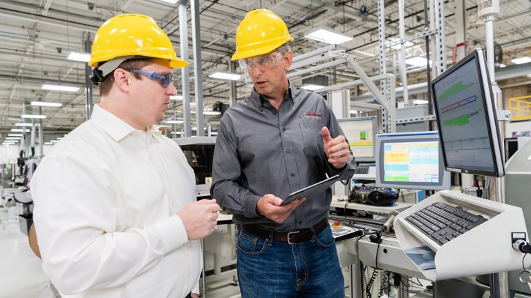 Two men in a factory both wearing yellow safety helmets and discussing what data on a tablet.