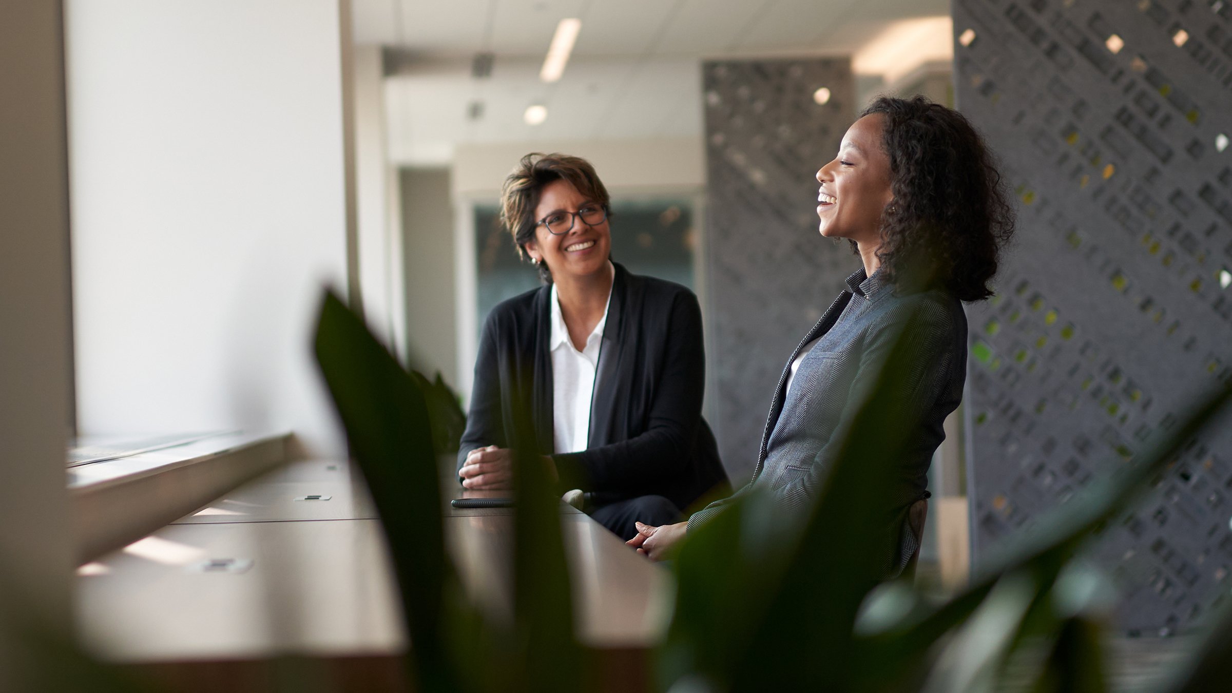 Two women sit at table by a window talking and smiling and laughing at Rockwell Automation Milwaukee headquarters