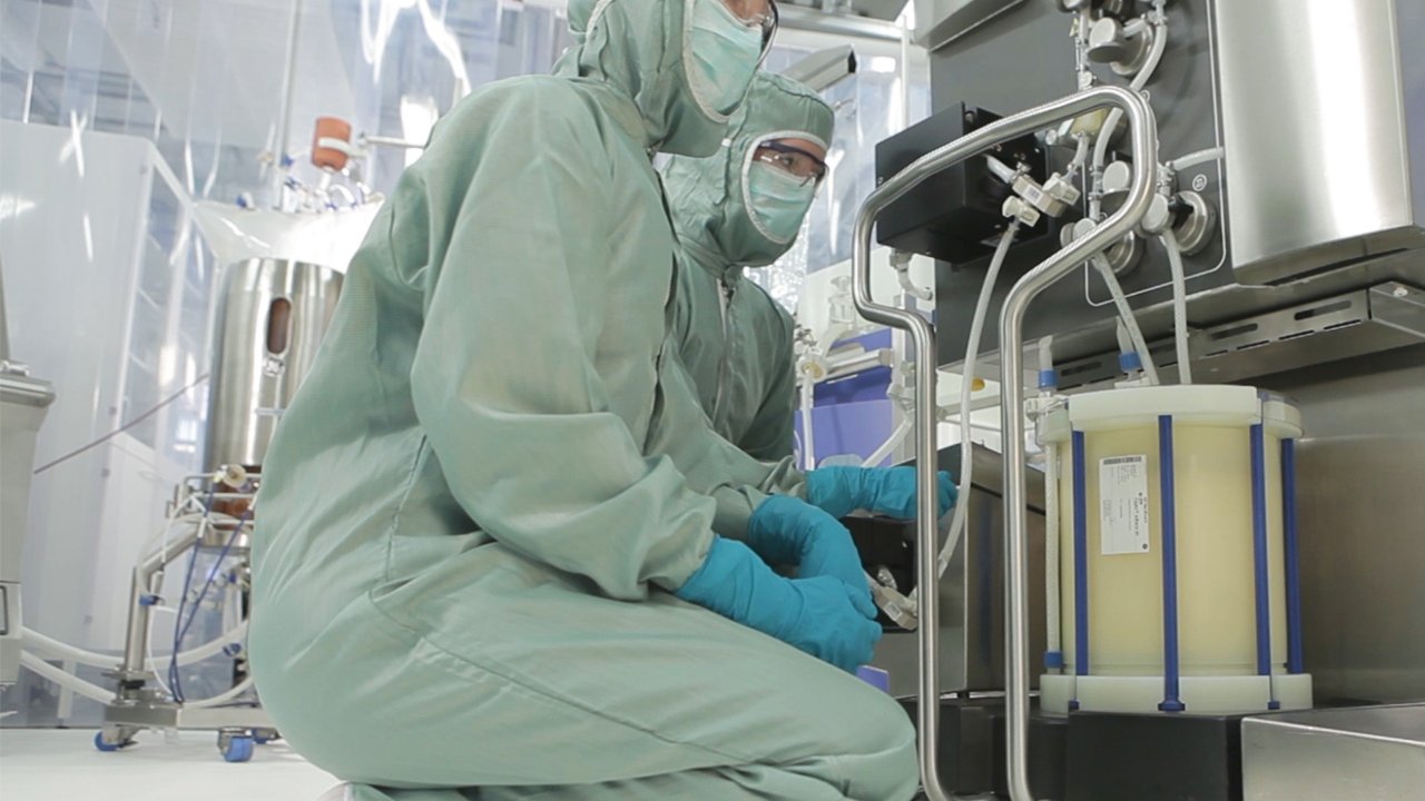 Two life sciences workers in full protective gear connect hoses to a single-use bioreactor bag in a manufacturing facility.