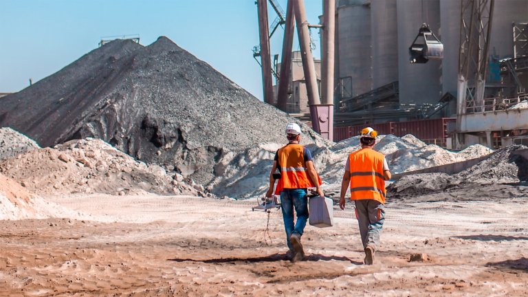 Two workers onsite at a cement production plant walking towards an area with heaps of raw materials that includes sand and soil