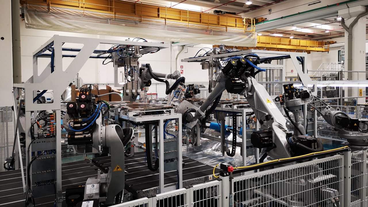 Cinco robôs Comau cinza de seis eixos em movimento enquanto operam em uma linha de produção de uma fábrica.