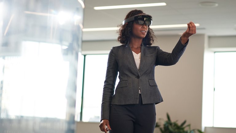 Una mujer en un ambiente de oficina moderno que lleva un casco de realidad aumentada y estira la mano en el aire como si quisiese tocar algo.