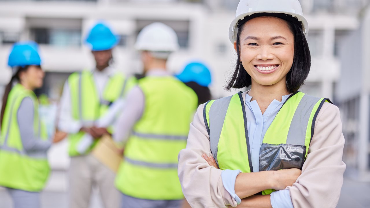 Asian female engineer crossed arm and wearing hard hat and safety apparel, smiling.