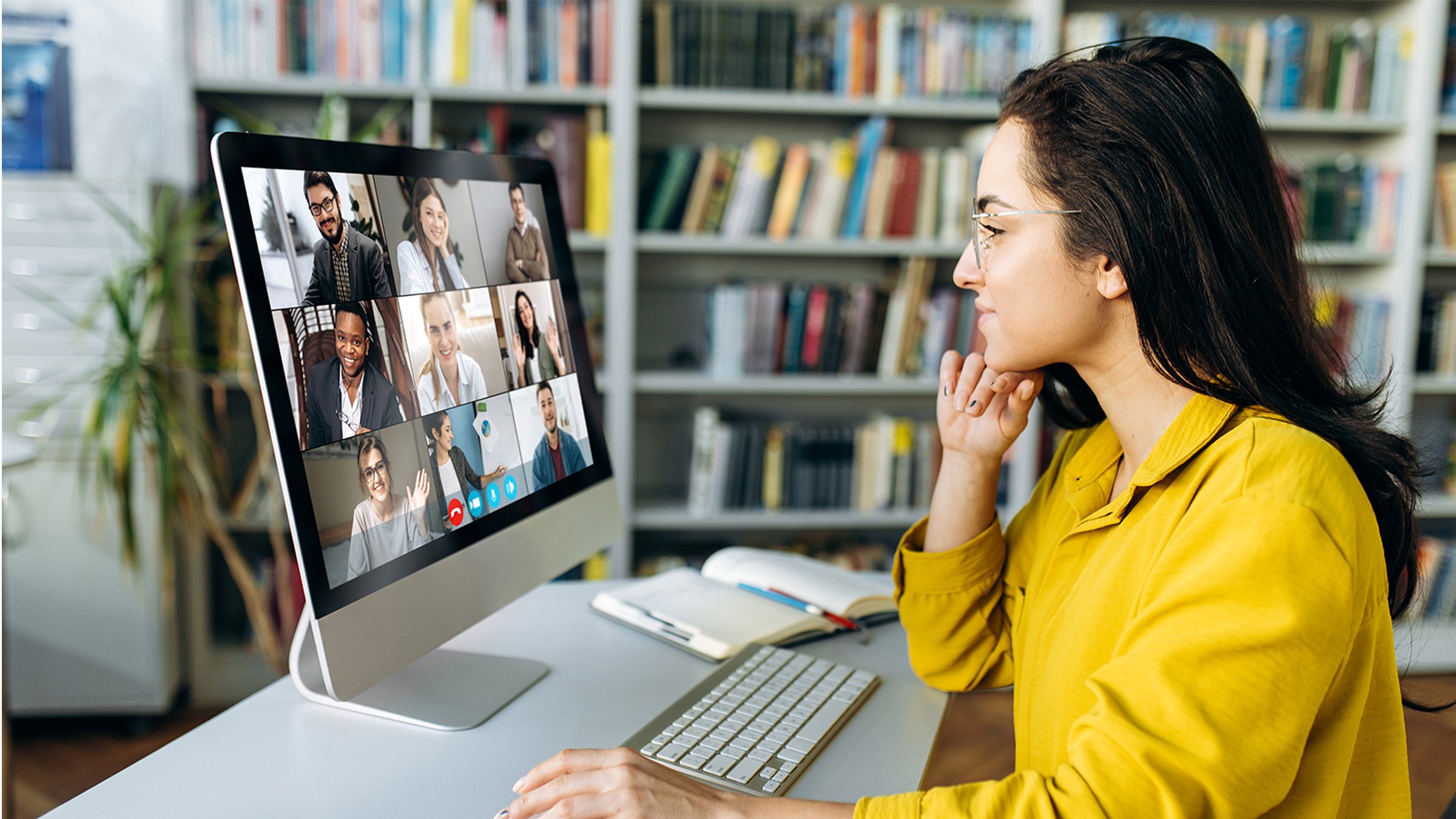 woman in yellow top doing on-line training in front of monitor with 9 individuals on monitor