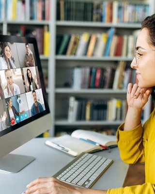 Frau mit gelber Bluse, die an einer Online-Schulung teilnimmt und auf einen Monitor blickt, auf dem 9 Personen zu sehen sind