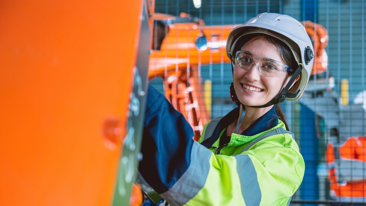 工廠中的年輕女性頭戴白色安全帽、佩戴護目鏡、身穿霓虹綠外套，並拿著筆電且檢查電子面板。在生產線中，橘色機械手臂出現在女性的身後。