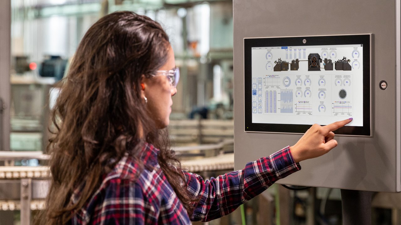 A woman wearing protective goggles in a factory setting uses a touchscreen panel to control industrial equipment