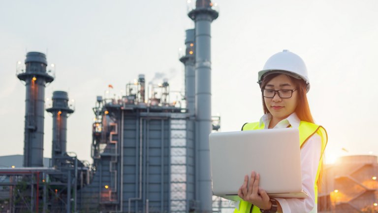 Woman wearing a hard hat and safety vest is holding a laptop, looking at the screen standing outside with a manufacturing plant behind her. 