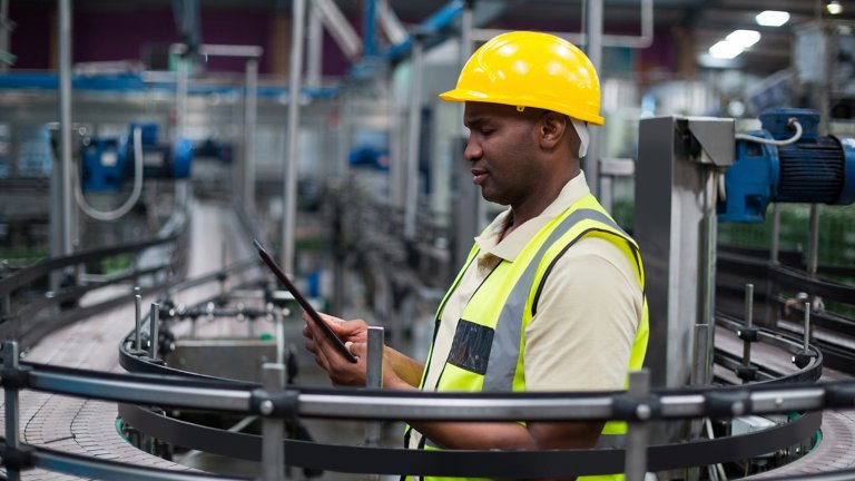 Pessoa com capacete e colete, olhando para o tablet entre máquinas no chão de produção