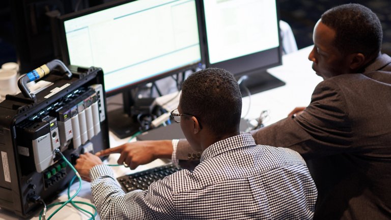 两名男子坐在计算机屏幕前参加 Rockwell Automation 在线学习课程并指向 ControlLogix 控制器。