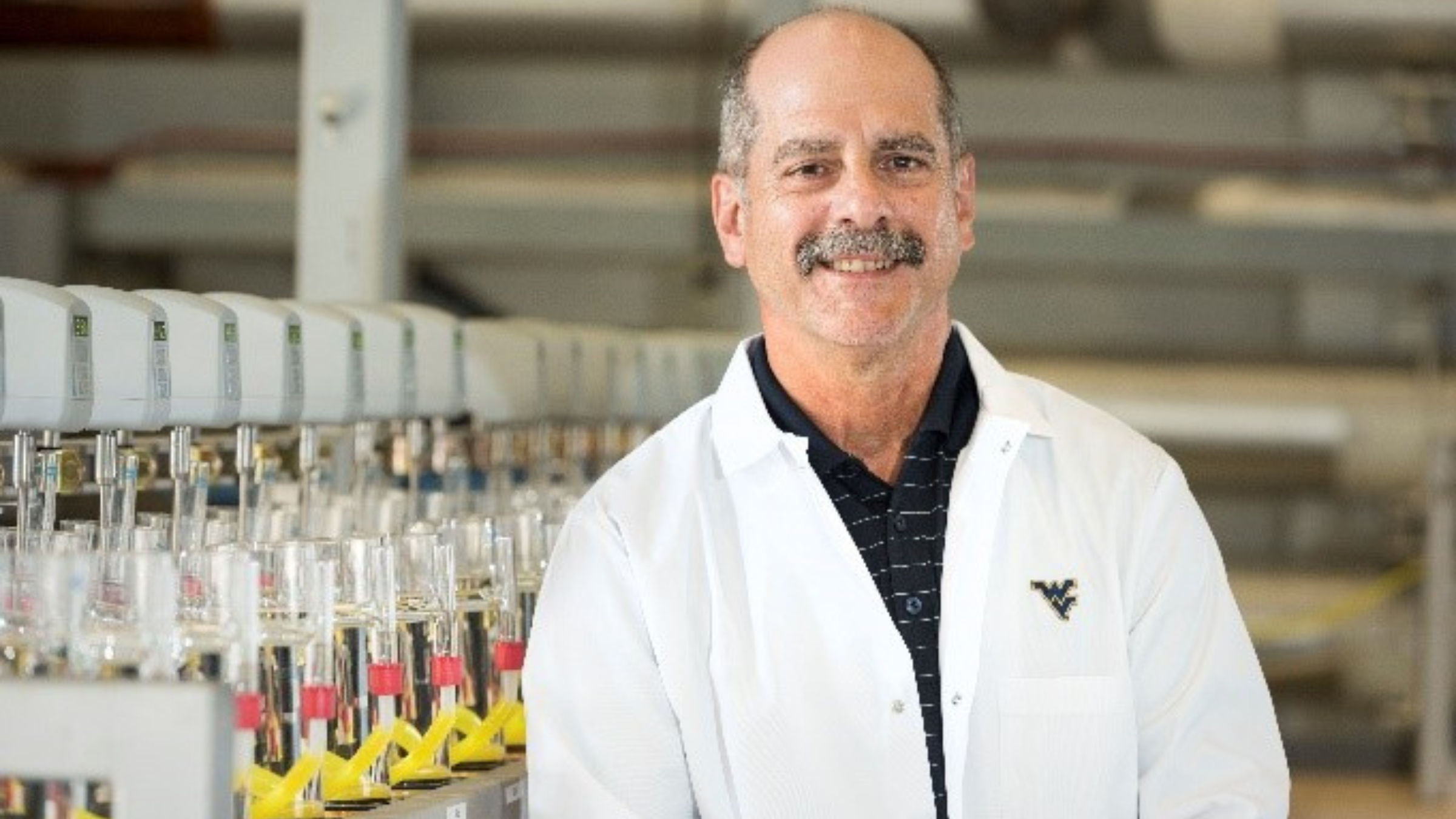 Male professor in white lab coat smiling in front of science lab equipment 