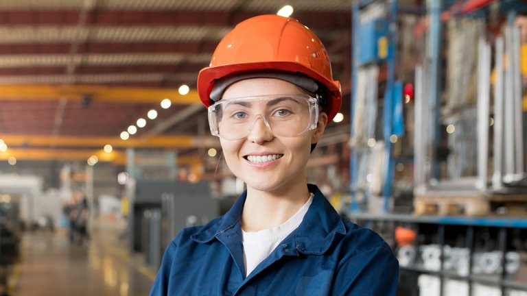 Young smiling female technician in blue uniform, protective eyeglasses and helmet working in modern factory