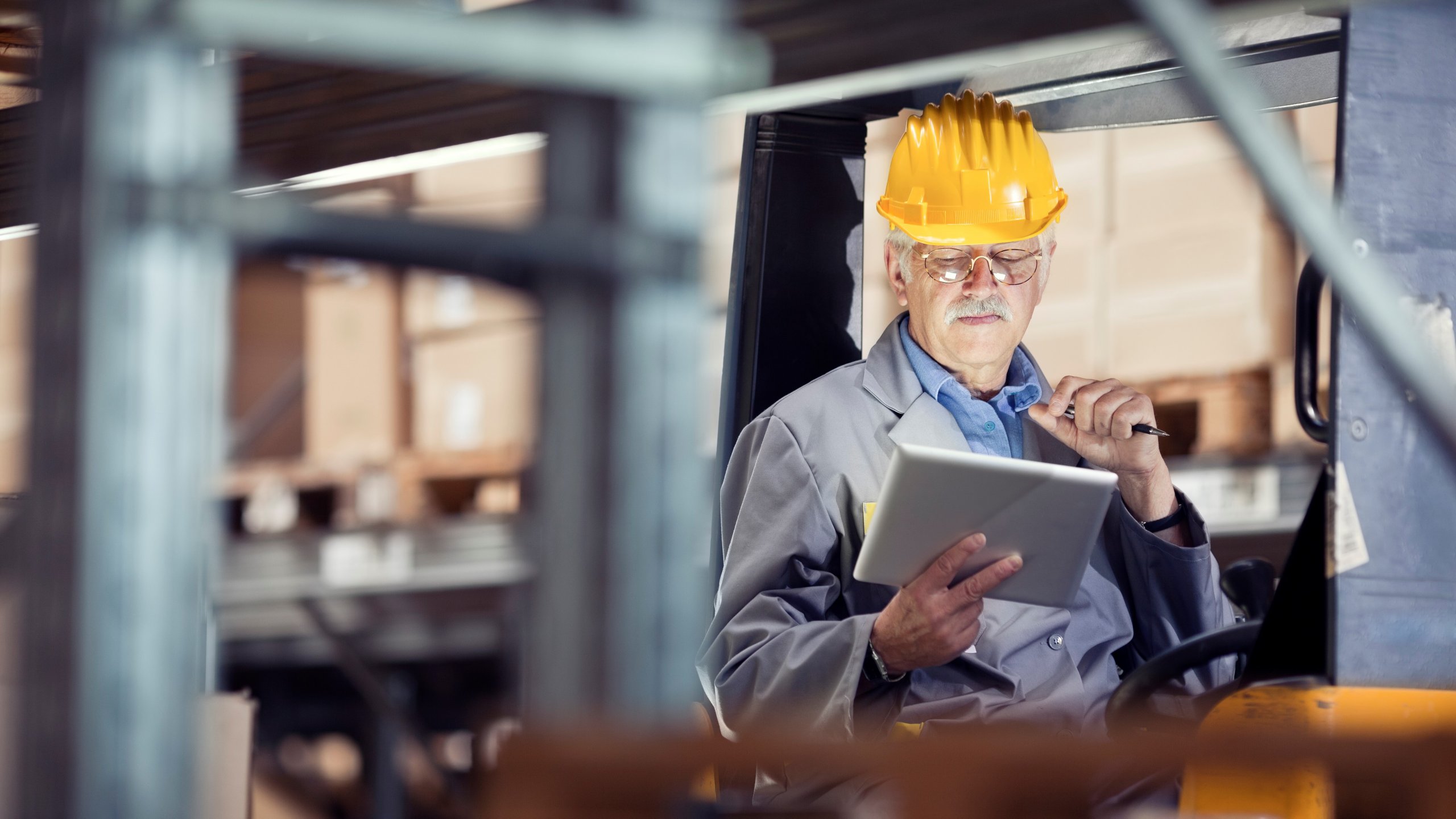 A man in a yellow hard hat standing near a manufacturing line and holding up a tablet de+AN48