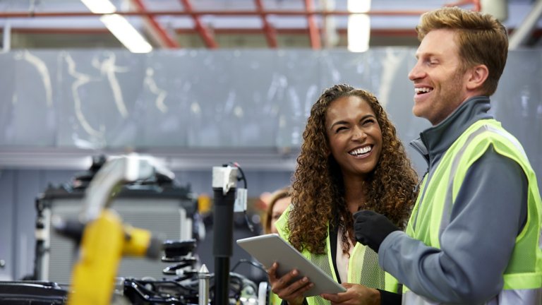 Two smiling engineers standing near manufacturing line and holding tablet