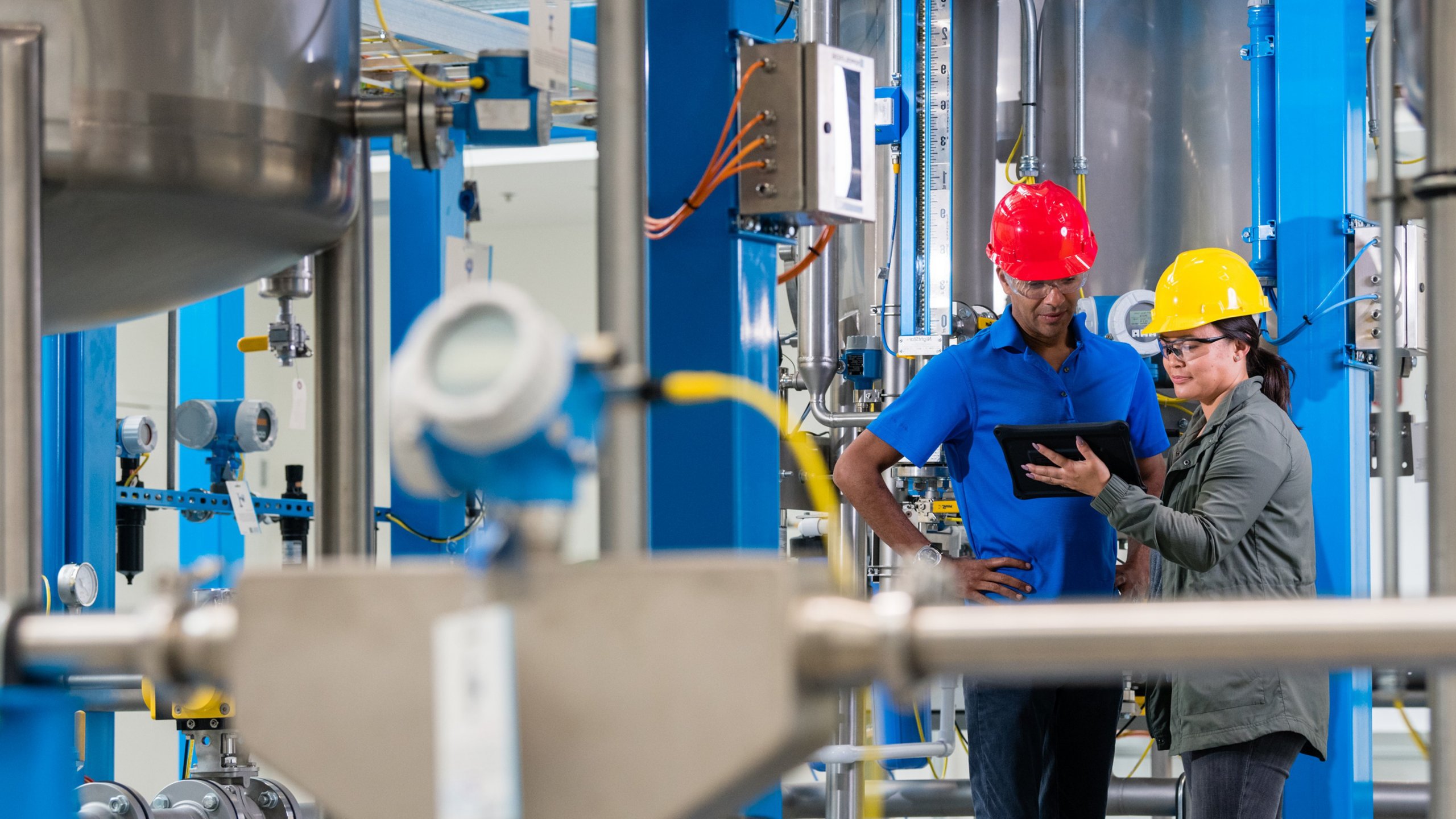 Um funcionário e uma funcionária em uma fábrica, usando capacetes e olhando para um tablet