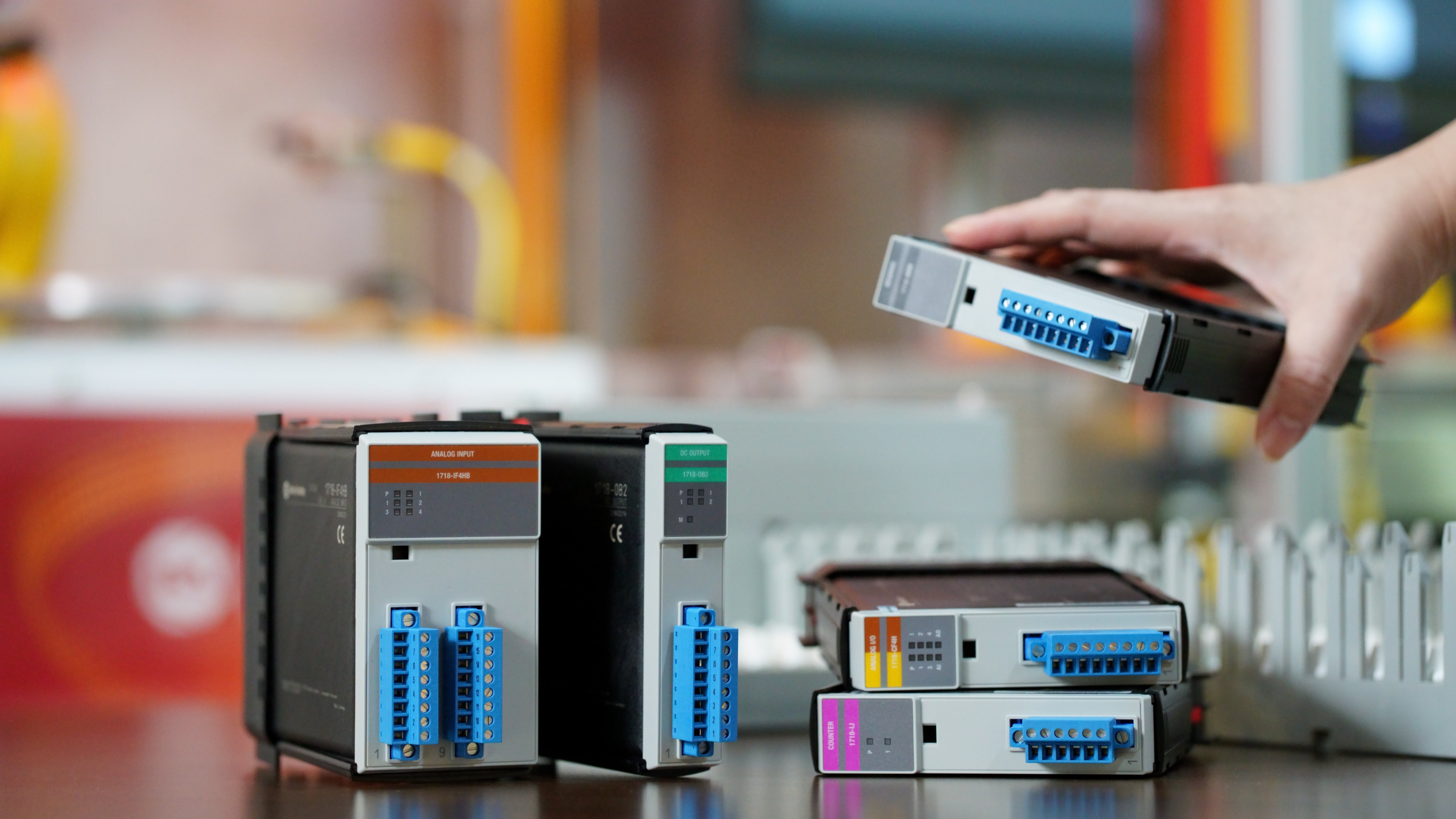 Front view image of five 1718 Ex I/O modules in a manufacturing plant environment. Four modules are places on a wooden table, one module is held by a person's hand at the middle right of the photo.