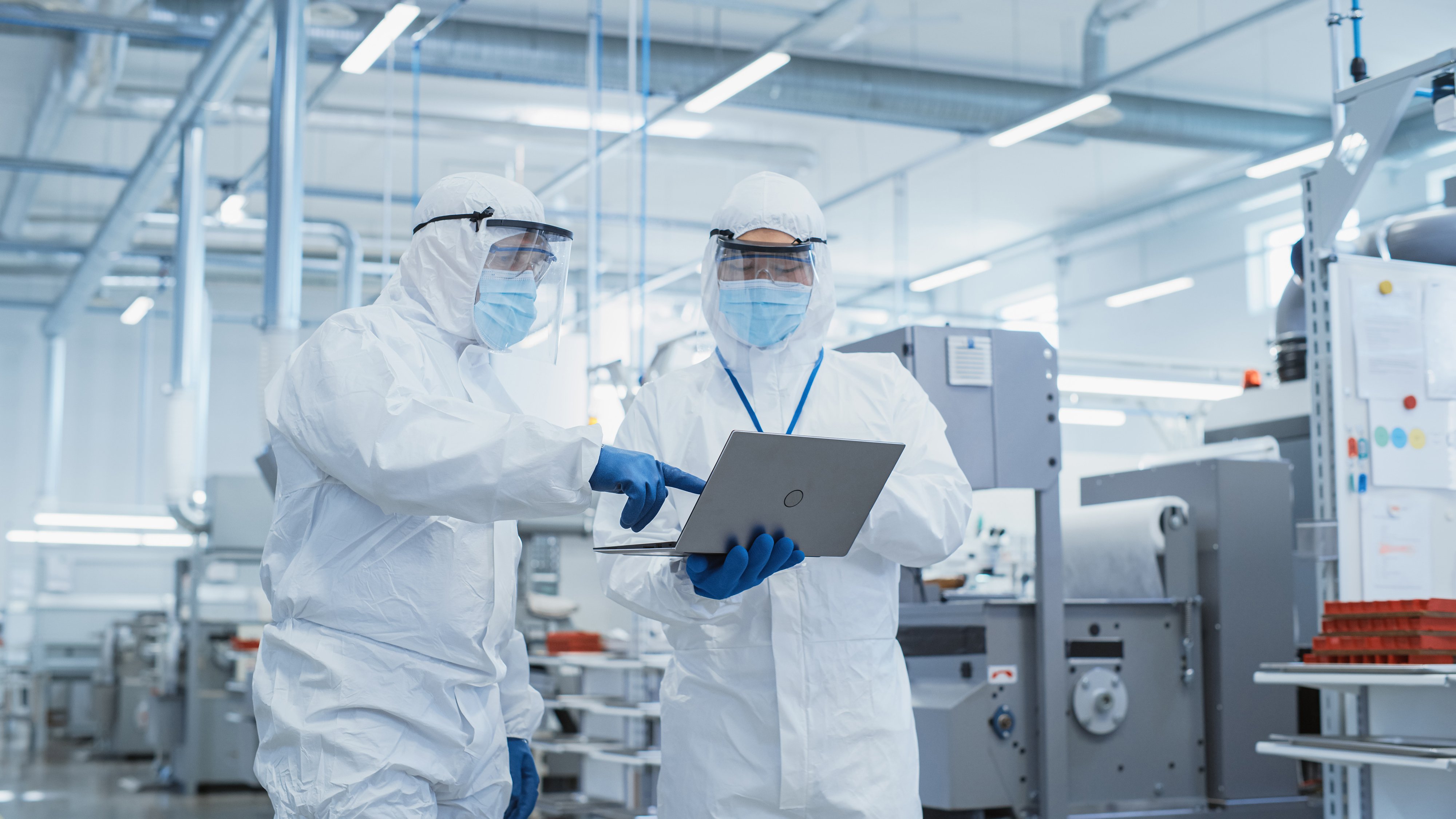 Two Scientists Walking in a Heavy Industry Factory in Sterile Coveralls and Face Masks, Examining data on laptop