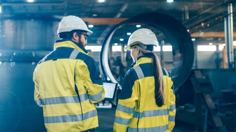 Dois trabalhadores usando capacetes e coletes amarelos em uma fábrica vendo informações em um tablet