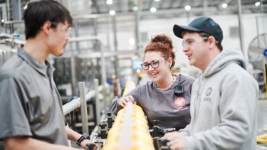 Three Church & Dwight employees, one female with red hair, two men, one wearing a blue baseball hat, all wearing protective eye gear, conversing on manufacturing production line. Conveyor belt is filled with yellow laundry detergent bottles.