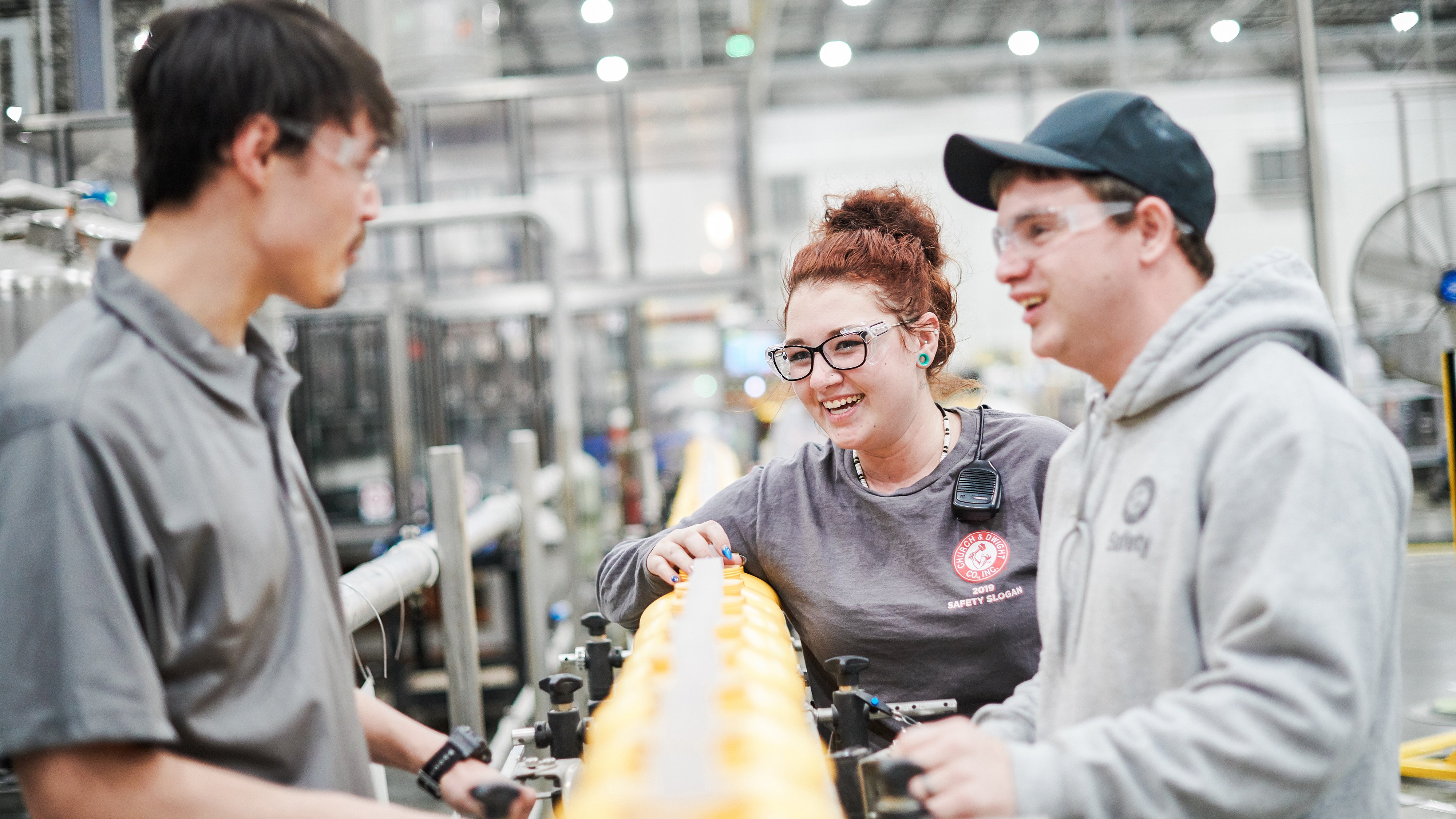 Three Church & Dwight employees, one female with red hair, two men, one wearing a blue baseball hat, all wearing protective eye gear, conversing on manufacturing production line. Conveyor belt is filled with yellow laundry detergent bottles. 