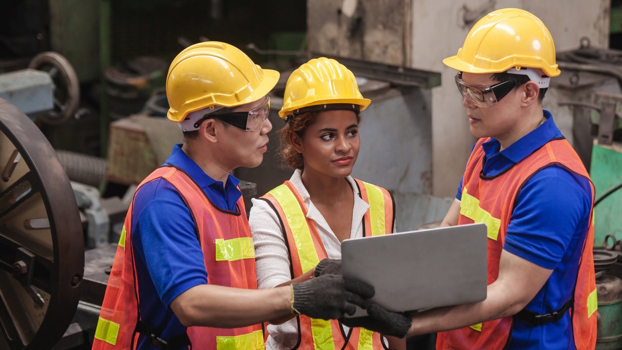 Three workers in PPE talk using laptop in factory