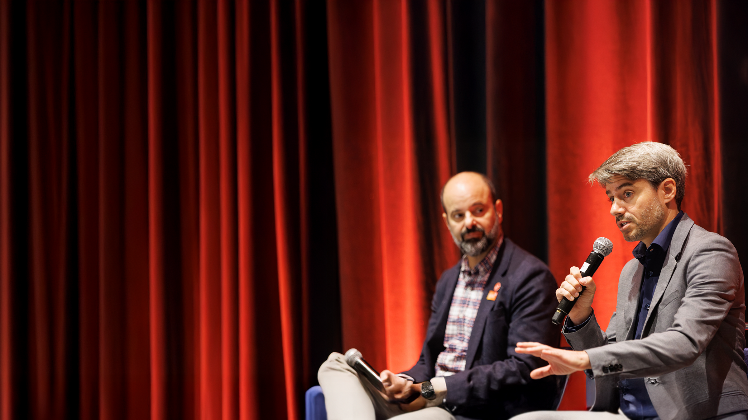 Two men sitting on a stage with microphones presenting