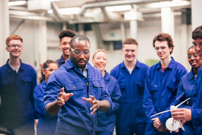 A tutor teaches his class about renewable energy in an engineering workshop. They are all wearing protective eyewear and blue coveralls.