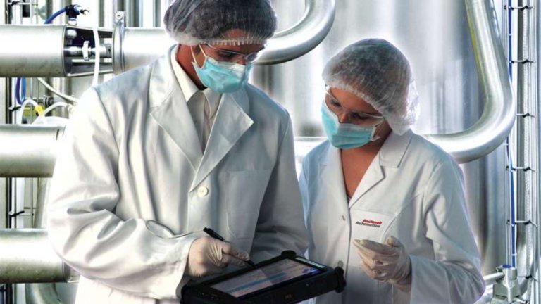 Two employees wearing hairnets, masks, white gloves and lab jackets viewing information on a tablet in a factory