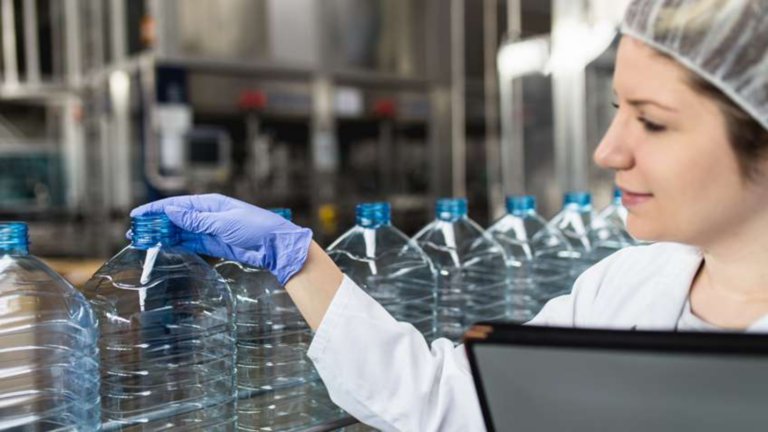 Junge glückliche Arbeiterin in einer Abfüllfabrik, die Wasserflaschen vor dem Versand kontrolliert. Überprüfung der Qualitätskontrolle.